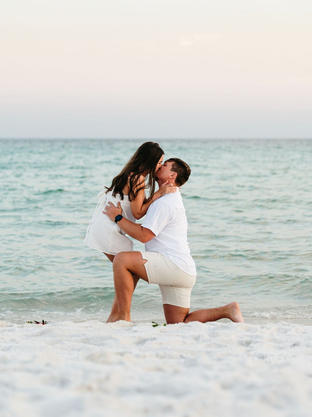 A couple embraces on a white sandy beach during a romantic seaside proposal at sunset.