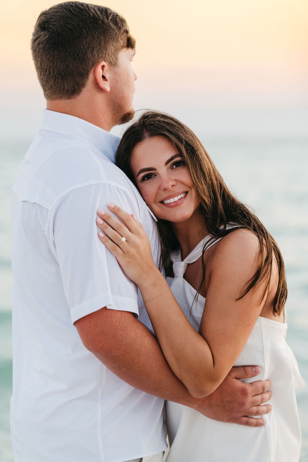 Couple embracing on a beach during sunset wearing white outfits.