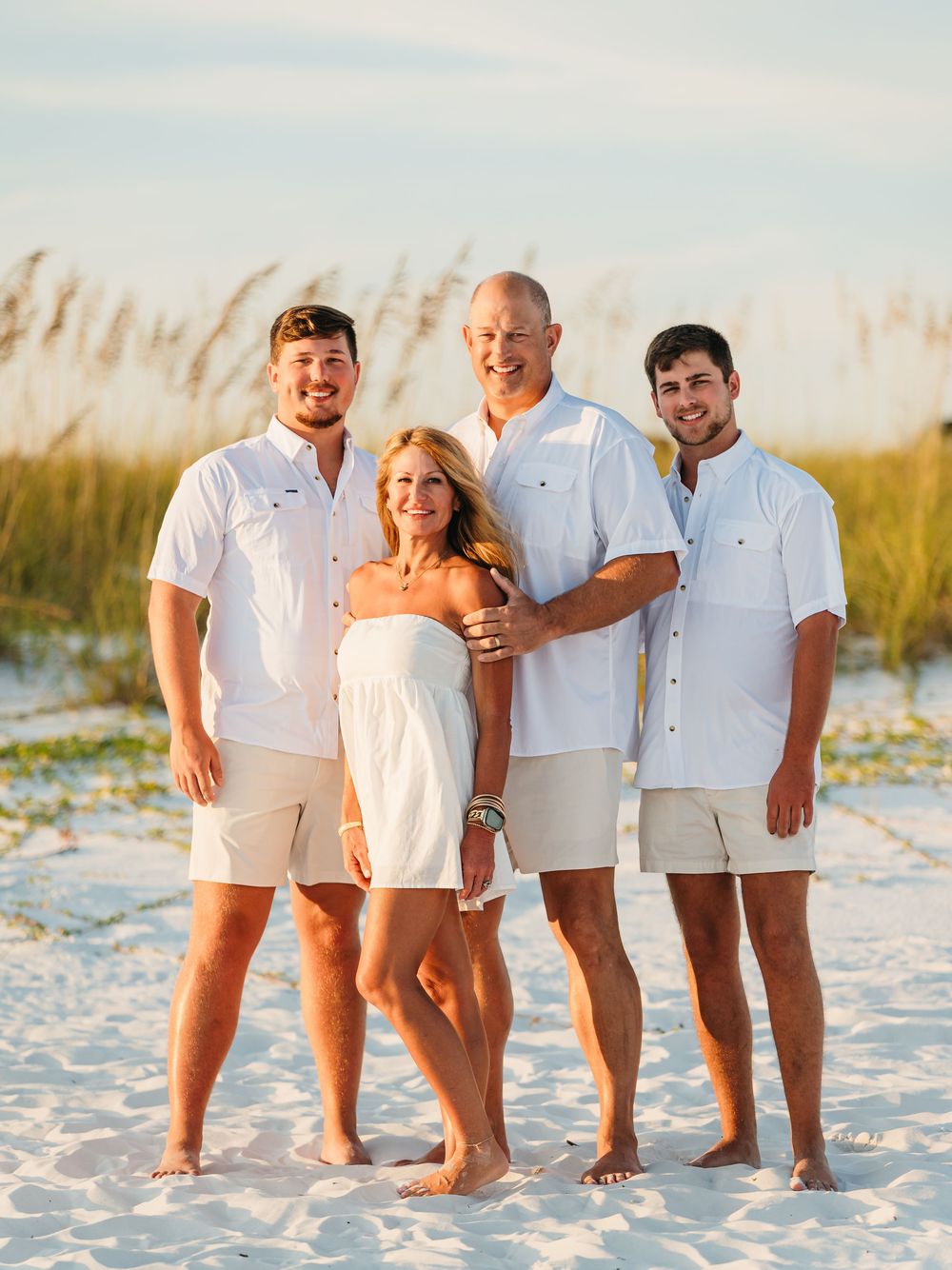 A group wearing white outfits stands together on a sandy beach at sunset with sea grass in the background.