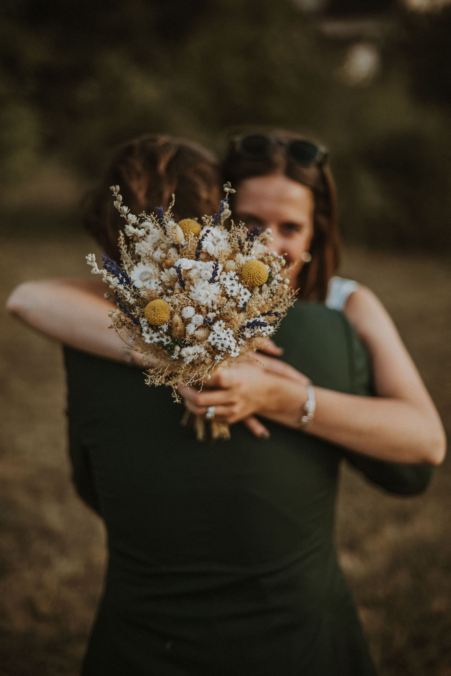 A dried floral bouquet with white blooms and yellow accents is held against a dark green background.