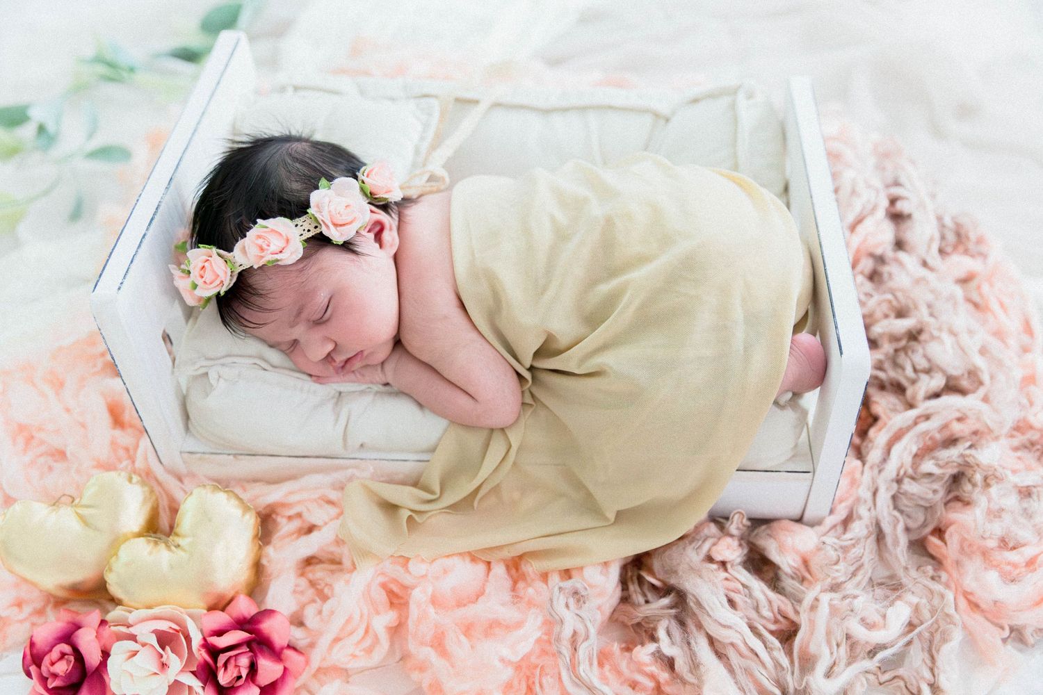 Sleeping newborn baby wrapped in yellow swaddle with floral headband in a white wooden mini crib.