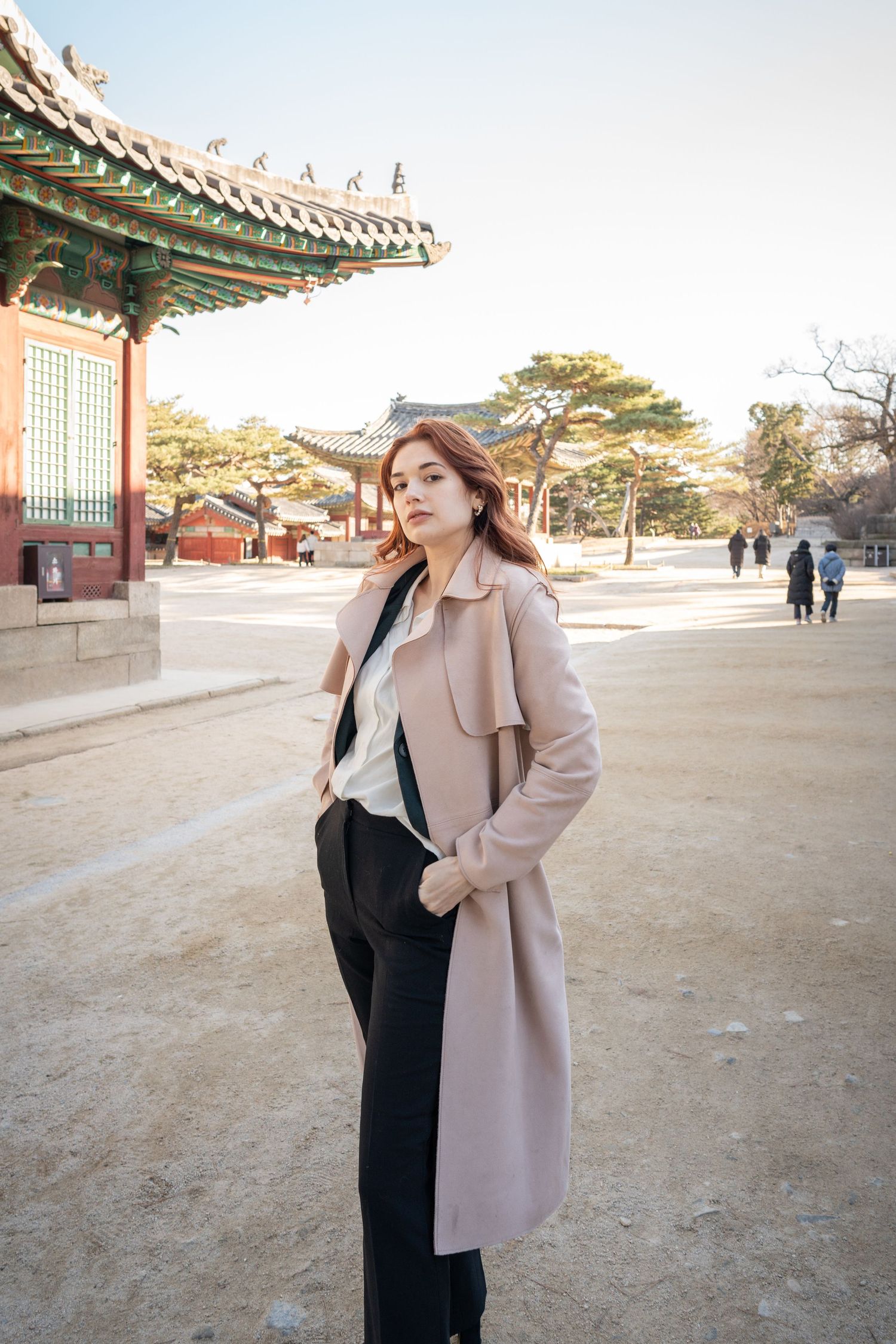 Stylish winter outfit with beige coat and black pants at traditional Korean palace courtyard with ornate architecture.