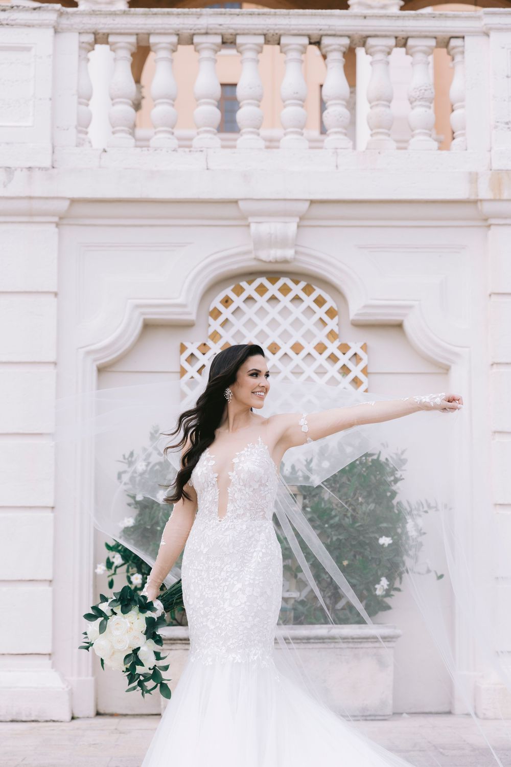 Bride in lace gown poses gracefully against white architectural backdrop.