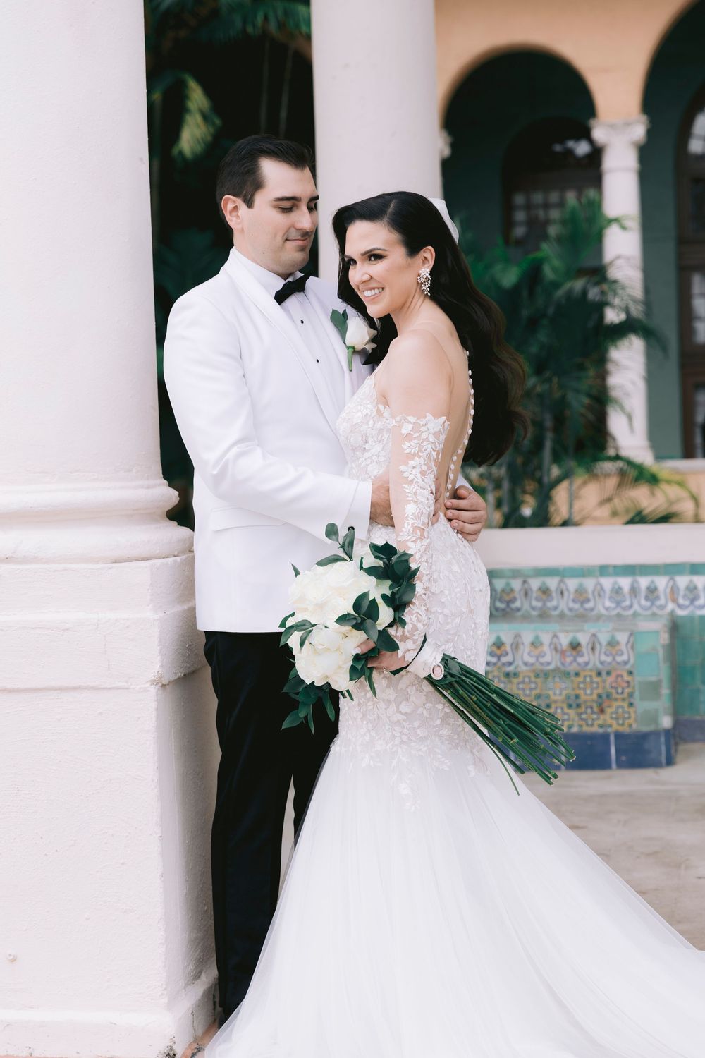 Elegant wedding couple poses against white columns with tropical foliage and decorative tiles in background.