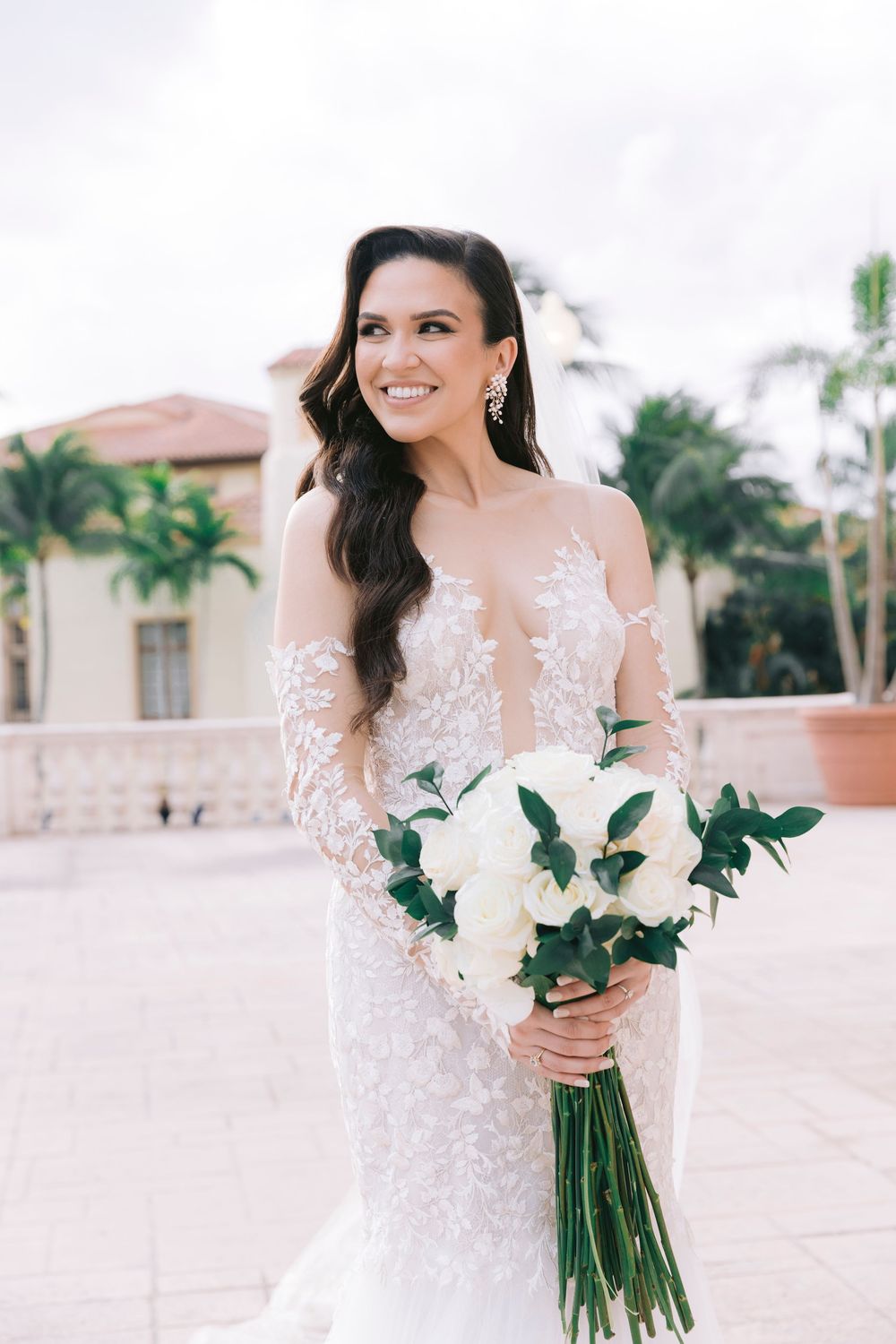Bride in lace detailed dress holds white floral bouquet against tropical resort backdrop.