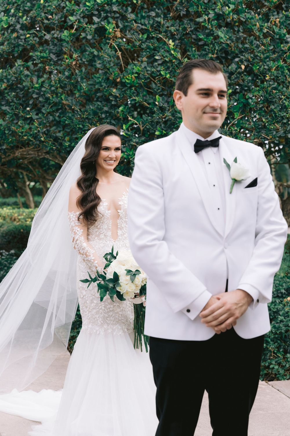 Wedding couple in white formal attire posing against green foliage backdrop.