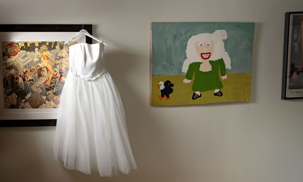 A white wedding dress hangs on display next to colorful artwork on a wall featuring a simple cartoon character.