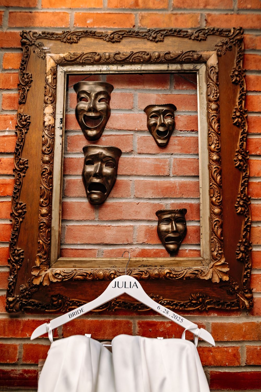 Ornate gold frames display theatrical comedy and tragedy masks mounted on a rustic brick wall.