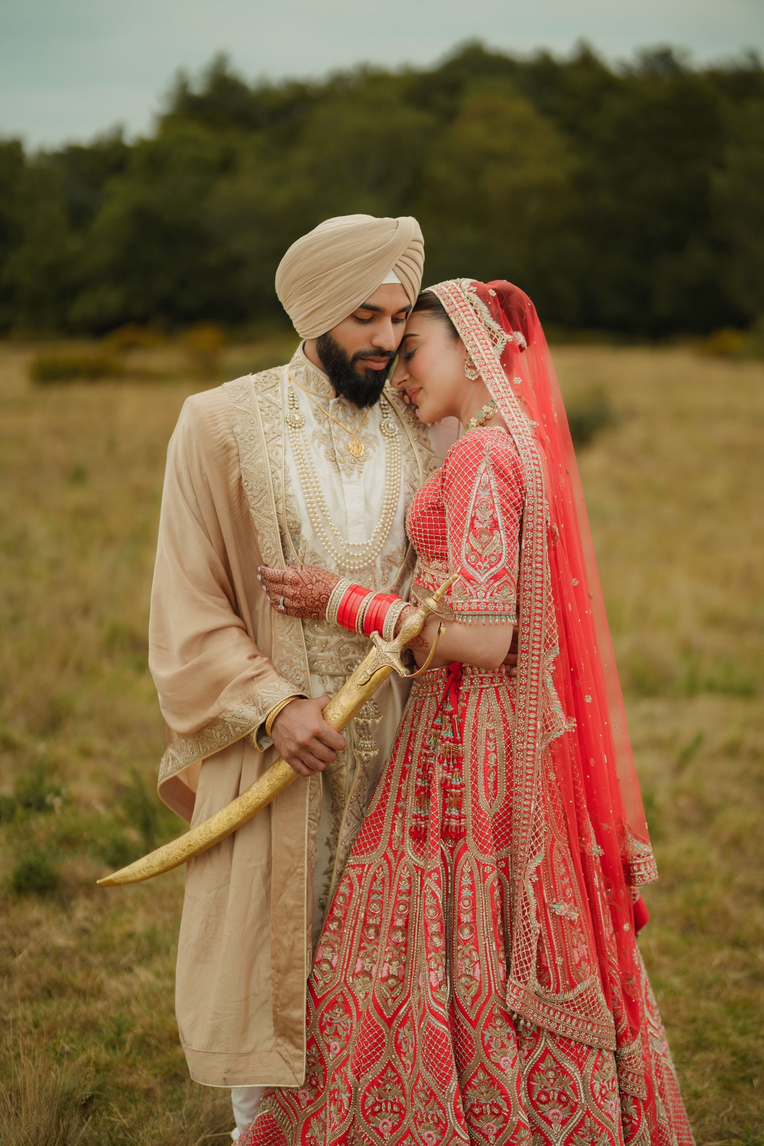 A couple in traditional Punjabi wedding attire poses together in a golden field during sunset.