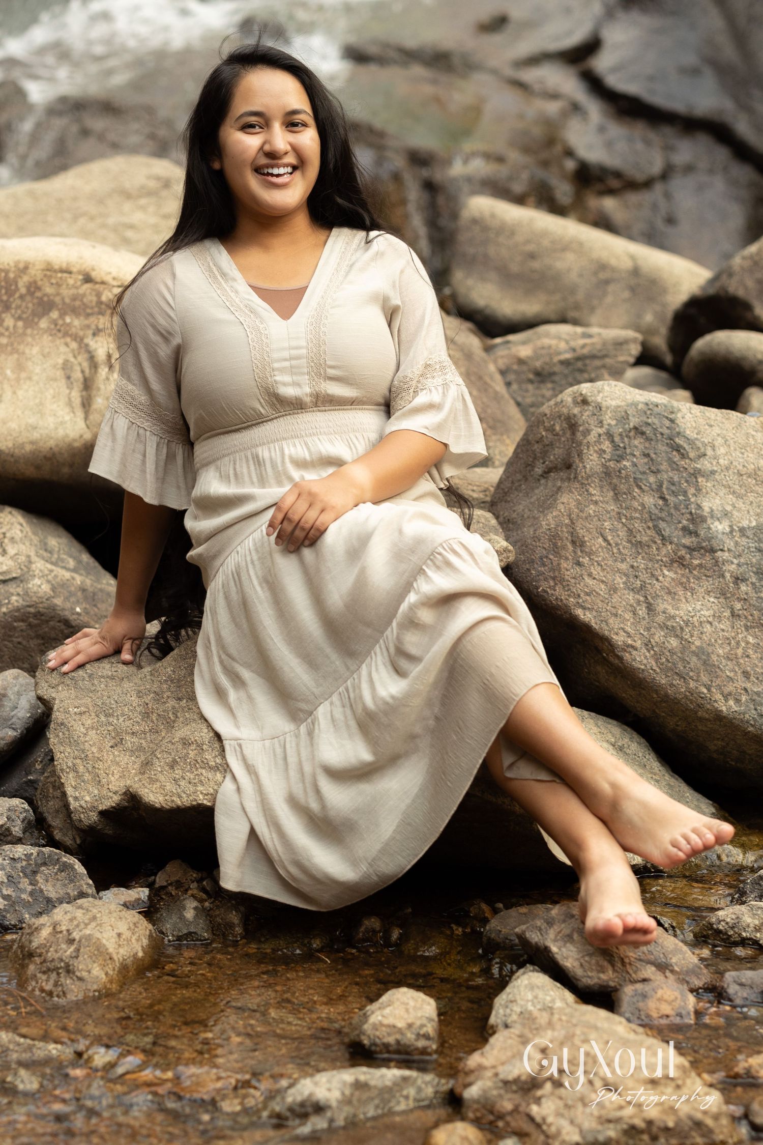 Person in flowing cream dress sitting barefoot on rocks by waterfall.