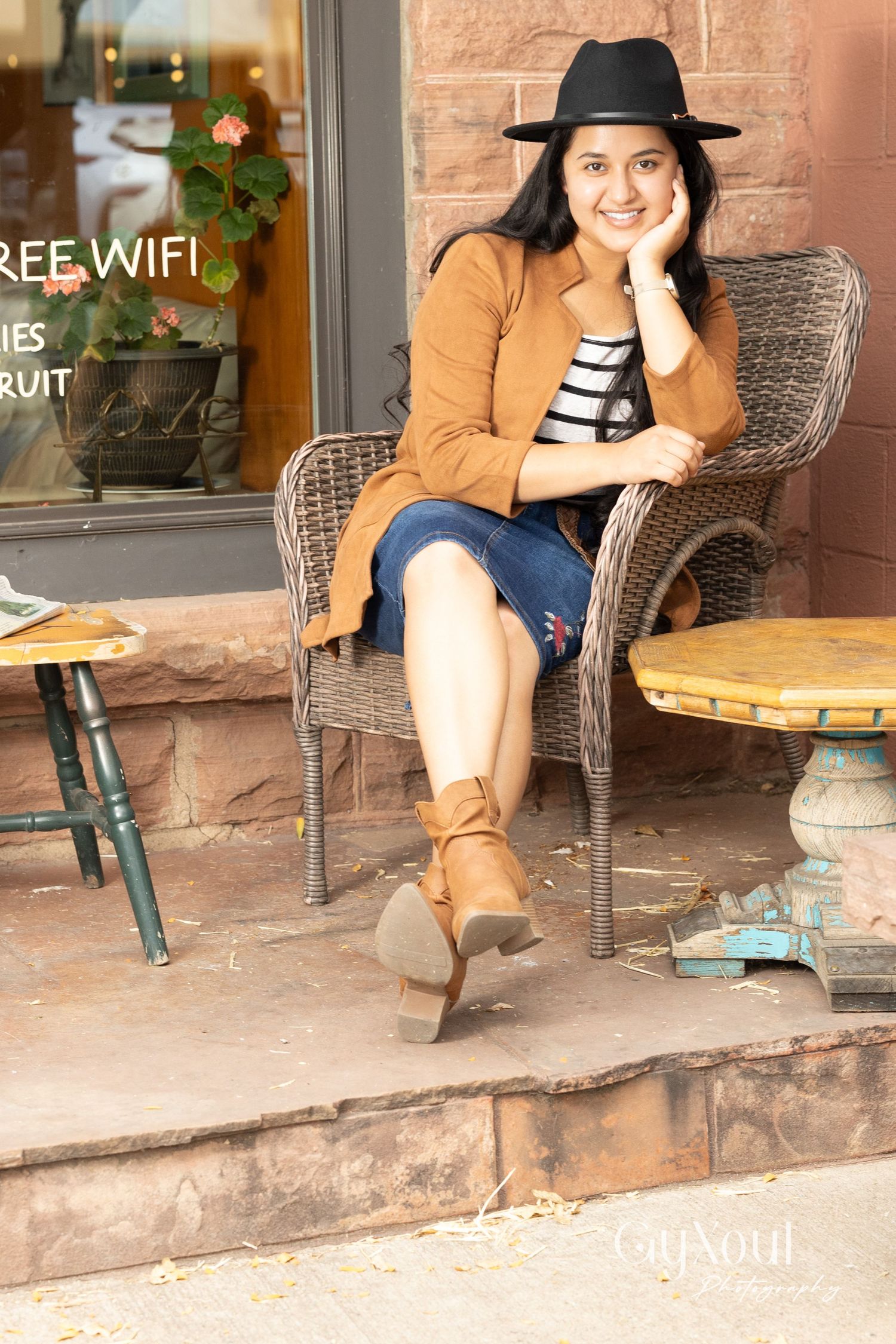 Person relaxing in wicker chair at cafe storefront wearing brown cardigan, striped shirt and black hat.