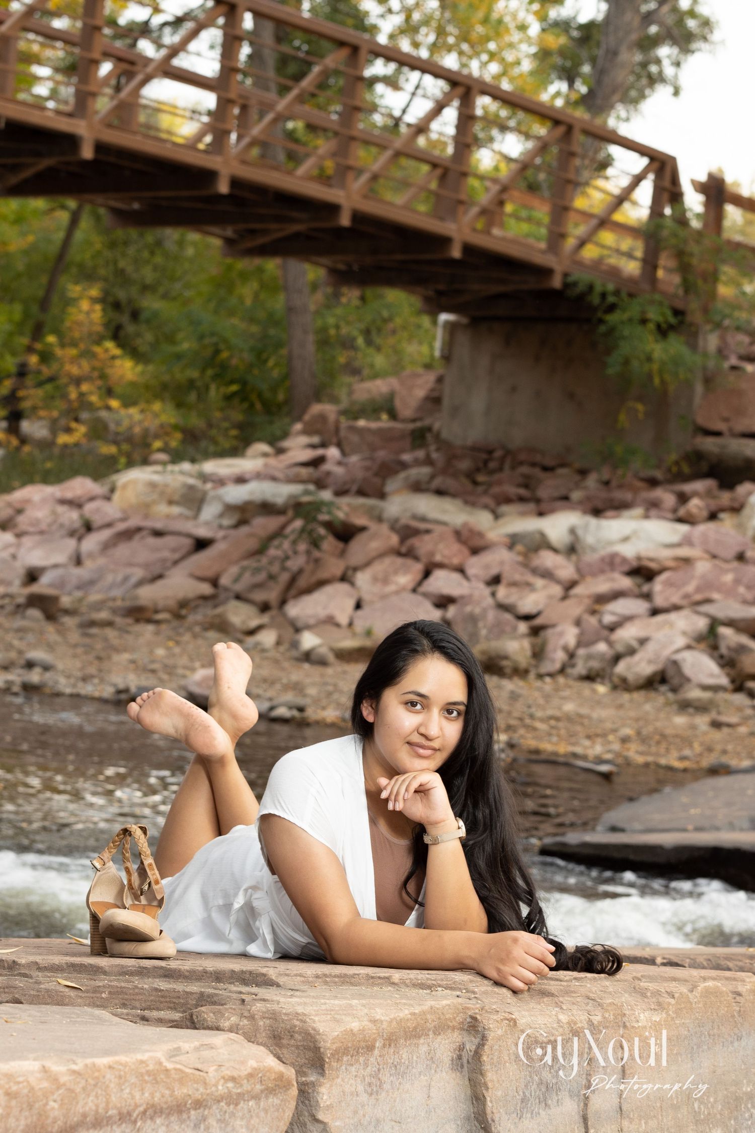 Person in white dress posing by rustic wooden bridge over rocky stream.