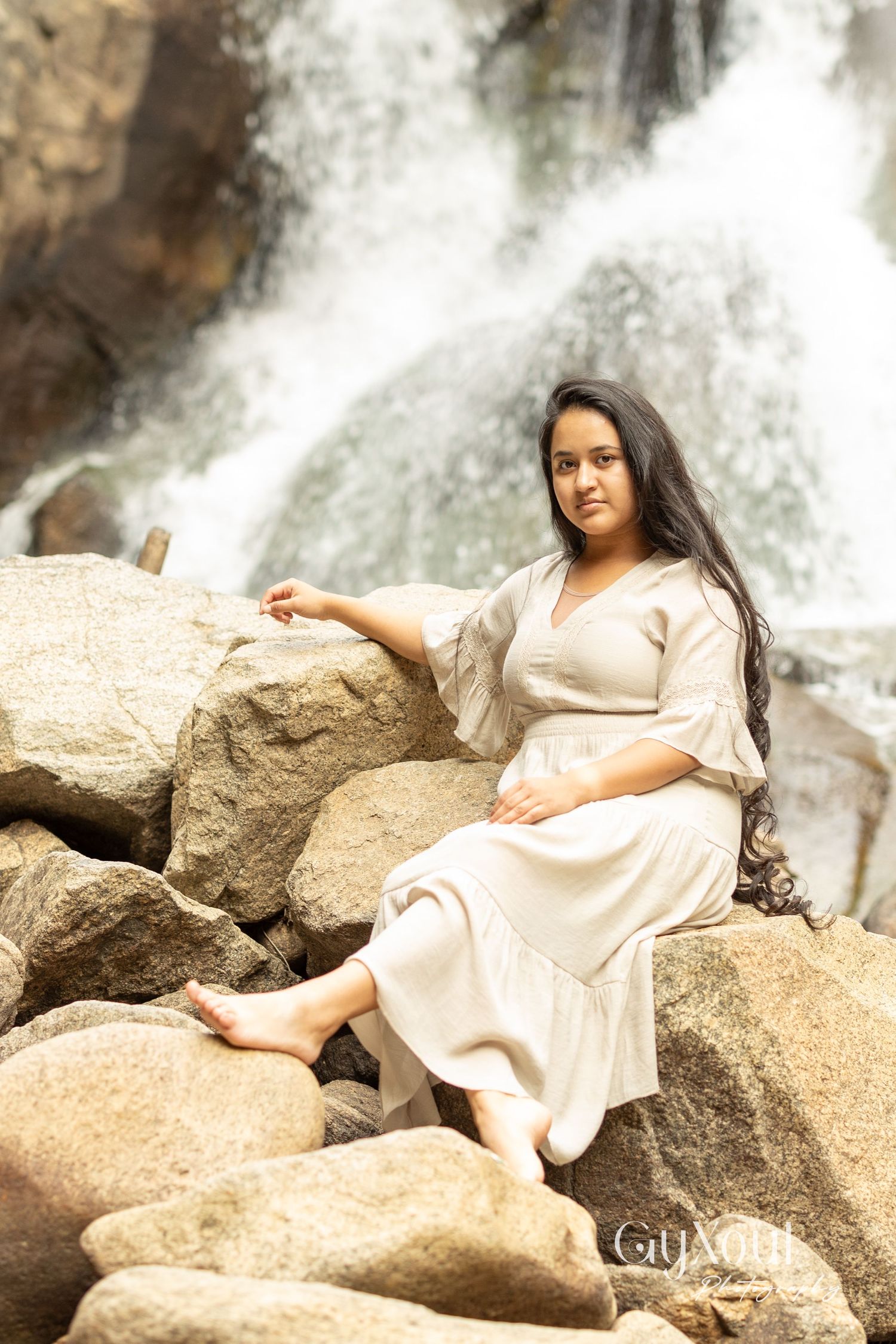 Person in beige dress sitting on large rocks near cascading waterfall.