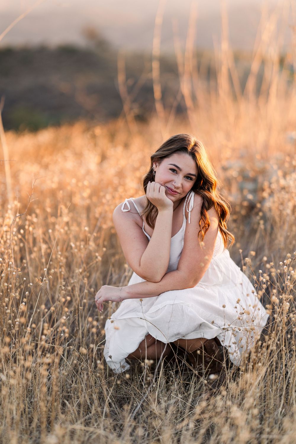 A woman in a white dress poses gracefully in a golden sunlit field of tall grass during sunset.