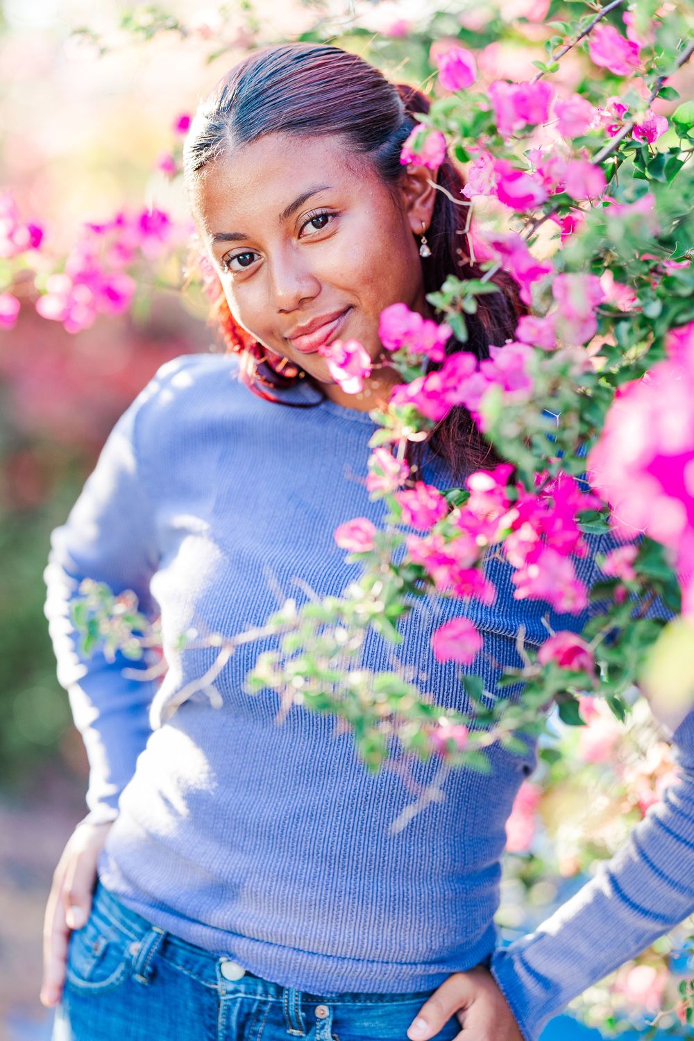 A person wearing a blue sweater poses among vibrant pink bougainvillea flowers in a garden setting.