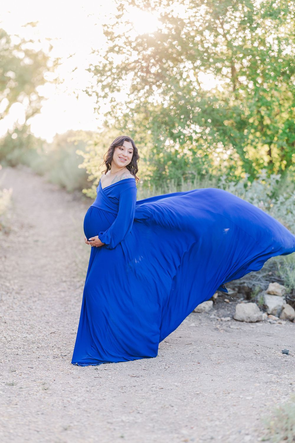 A woman in a flowing royal blue dress poses gracefully on the ground in a natural outdoor setting during golden hour.