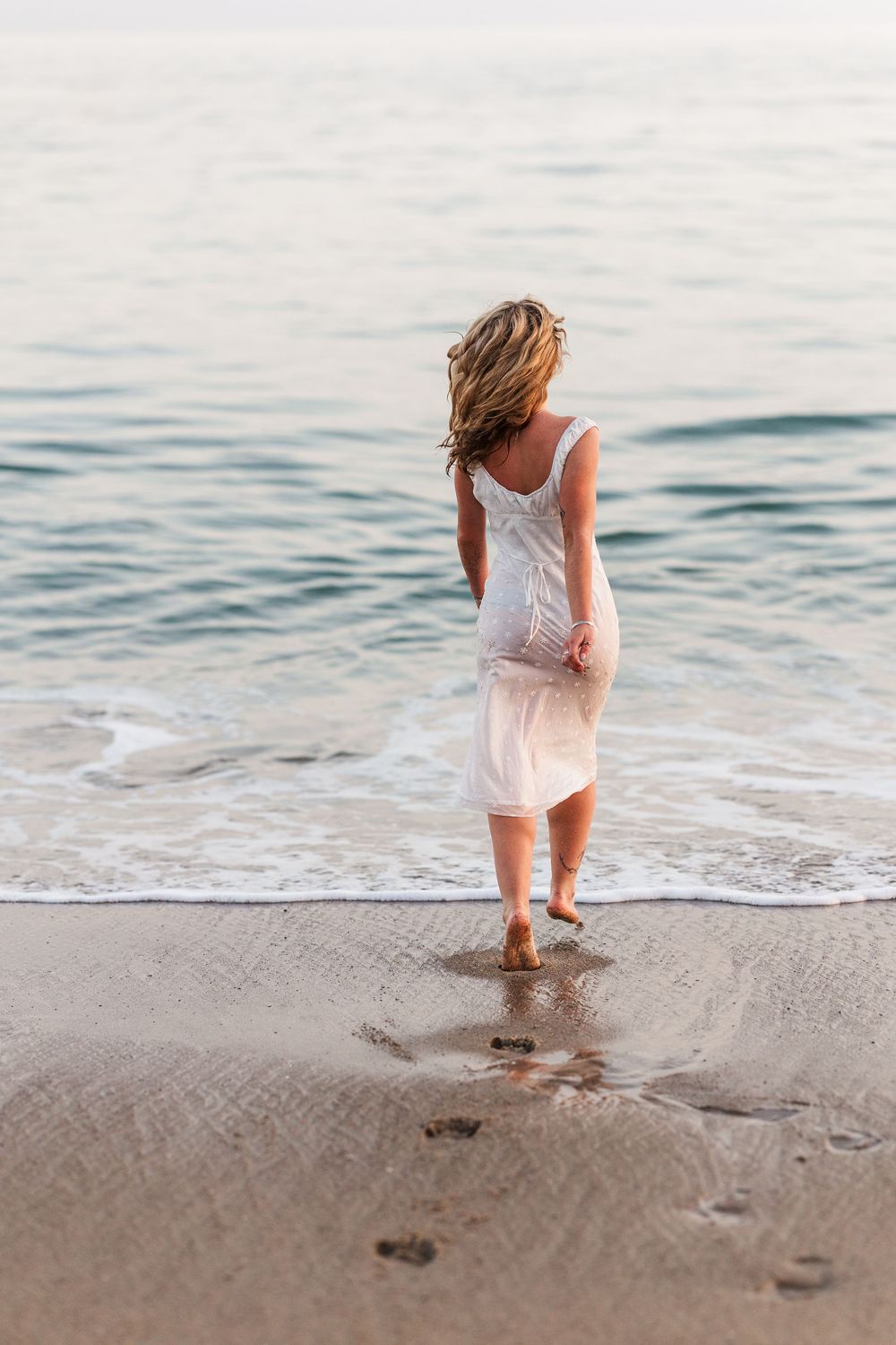 A person in a white dress walks along the beach shoreline leaving footprints in the sand at sunset.