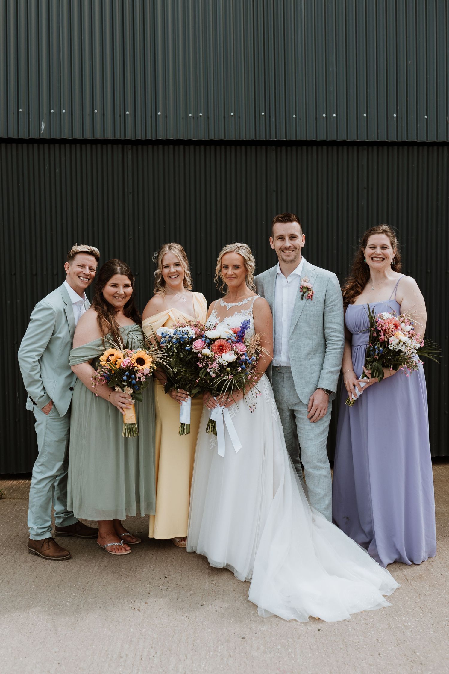 Wedding party poses together in front of green wall wearing mint suits and lavender bridesmaid dresses at Stanford Barn