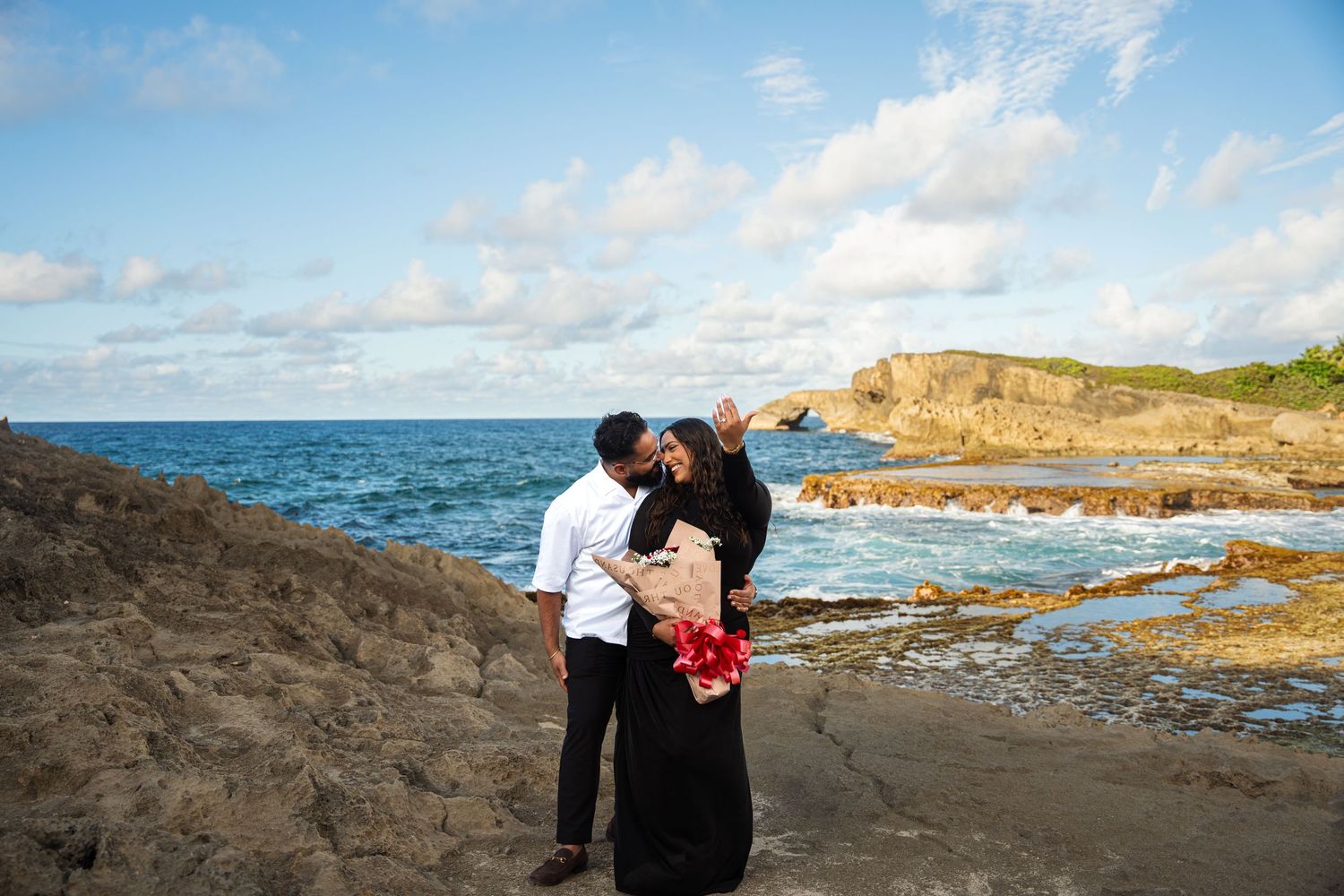 Couple embraces on a rocky coastal cliff with dramatic ocean views and cloudy skies in the background.