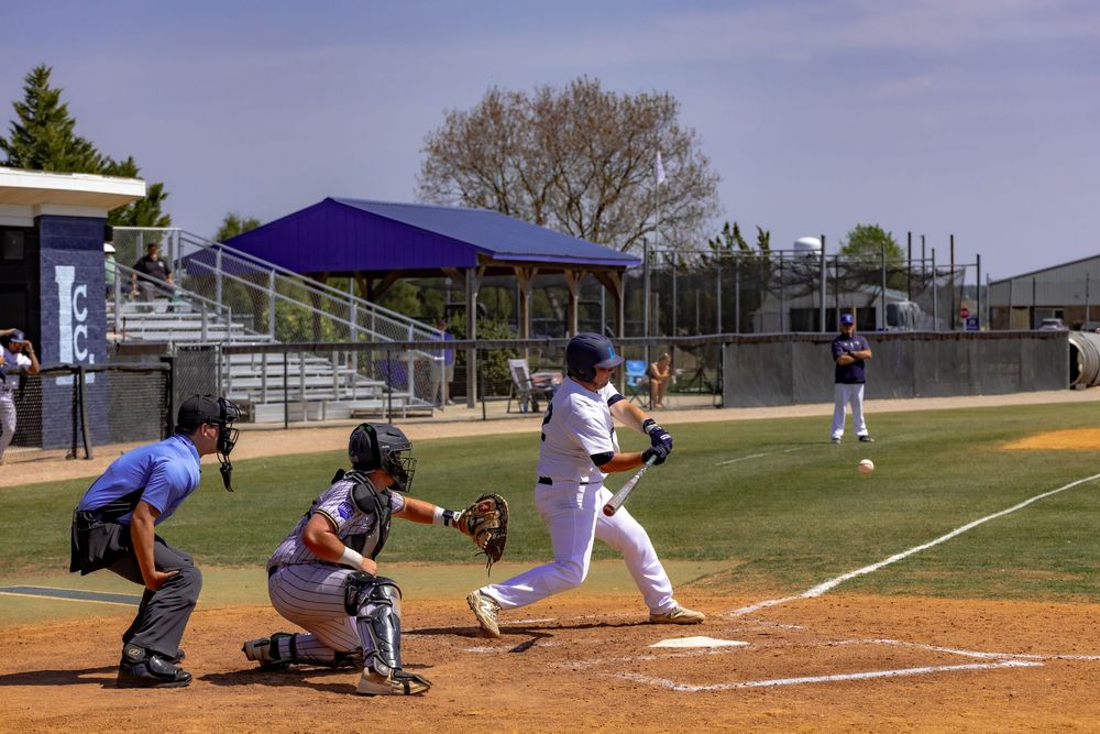 Baseball batter swings at pitch as catcher and umpire watch during a sunny outdoor game on a dirt infield.