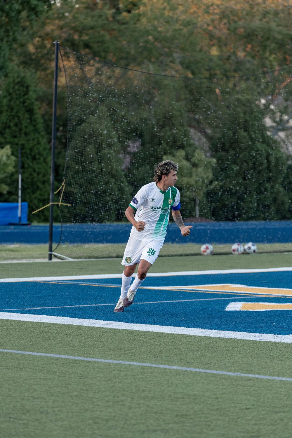 Soccer player in white and green uniform running on blue turf field during a rainy match.