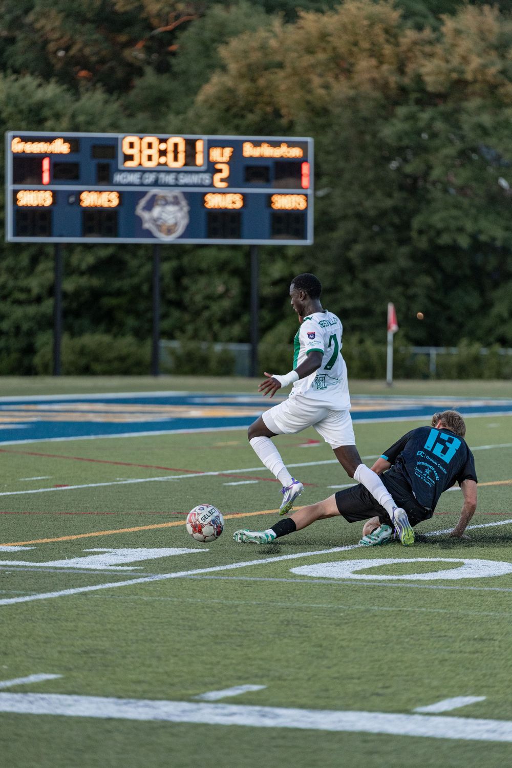 Soccer players compete for the ball during a night match with scoreboard showing 98:01 in the background.