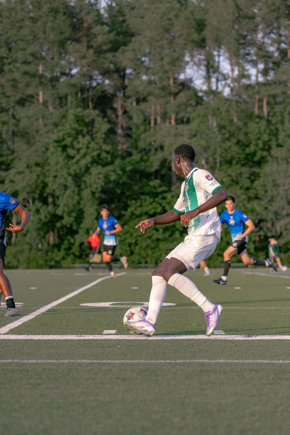 Young soccer player in white and green uniform dribbling ball on turf field during competitive match with trees in background.