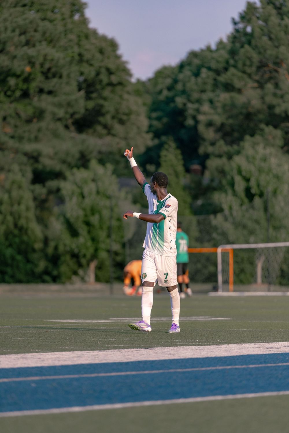 Soccer player in white jersey raises hand on blue track field with green trees in background.