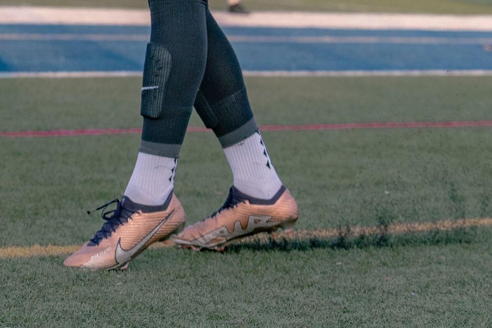 Close-up of athlete's legs wearing gold Nike cleats and white socks on a green turf field.