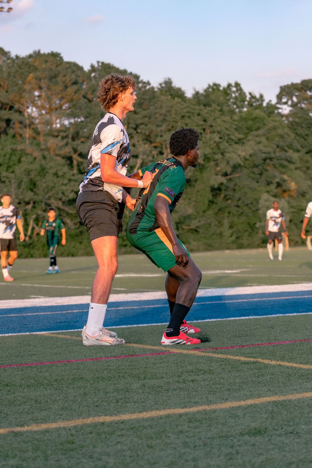 Two soccer players compete on a blue-lined field, one jumping in a green uniform, the other in white, with trees in background.
