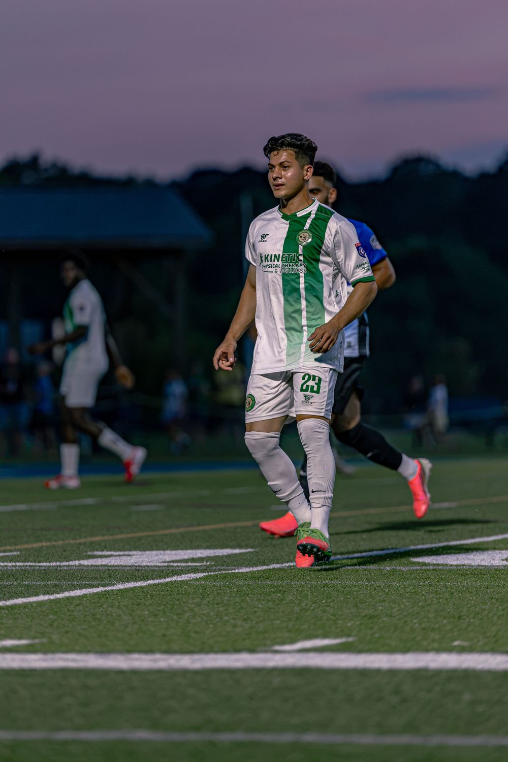 Soccer player in white and green uniform dribbling on field at dusk during a competitive match.