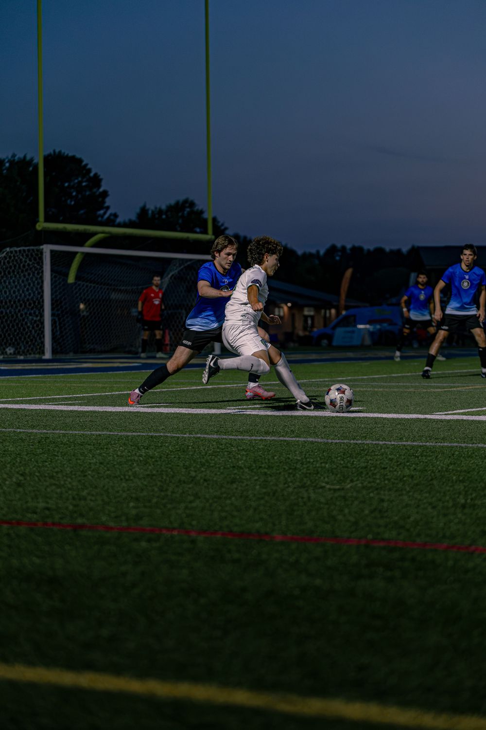 Two soccer players compete for the ball on a turf field under evening lights during a competitive match.