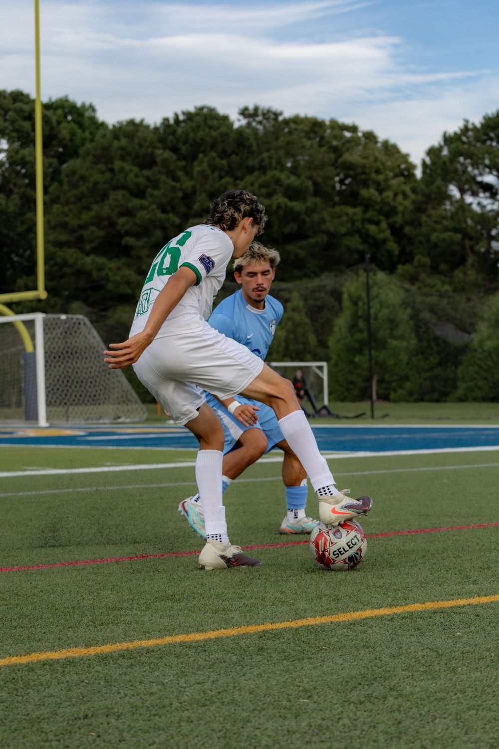 Two soccer players compete for the ball during a match on a turf field, one in white and one in light blue jersey.