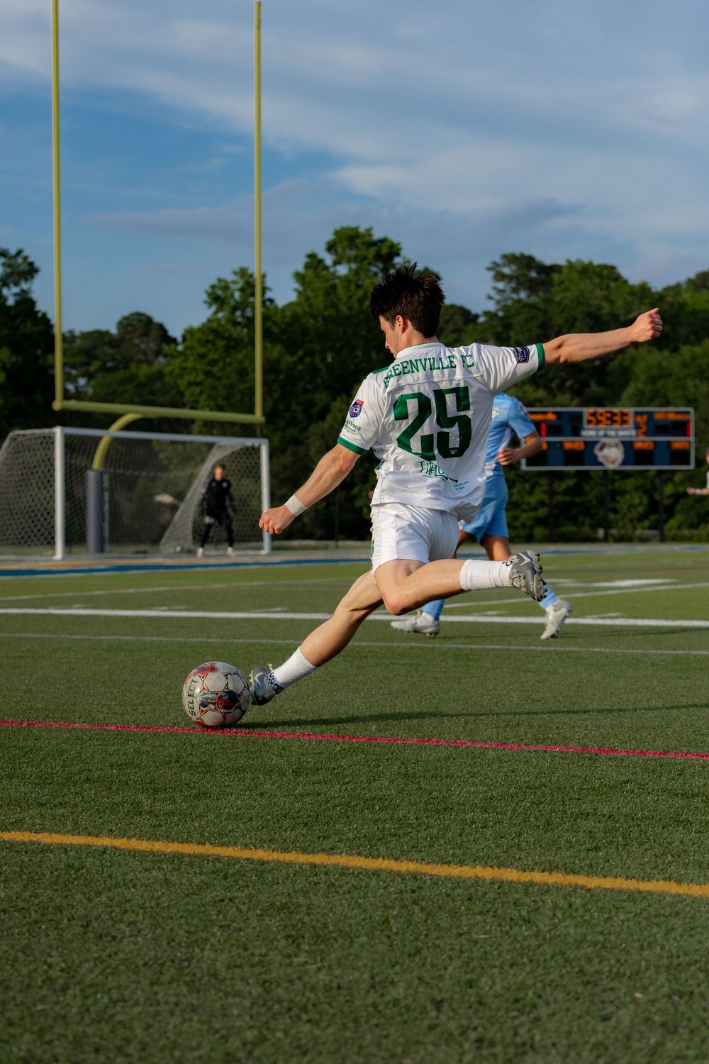 Soccer player wearing jersey number 25 kicking a ball on a turf field during a game under a blue sky.