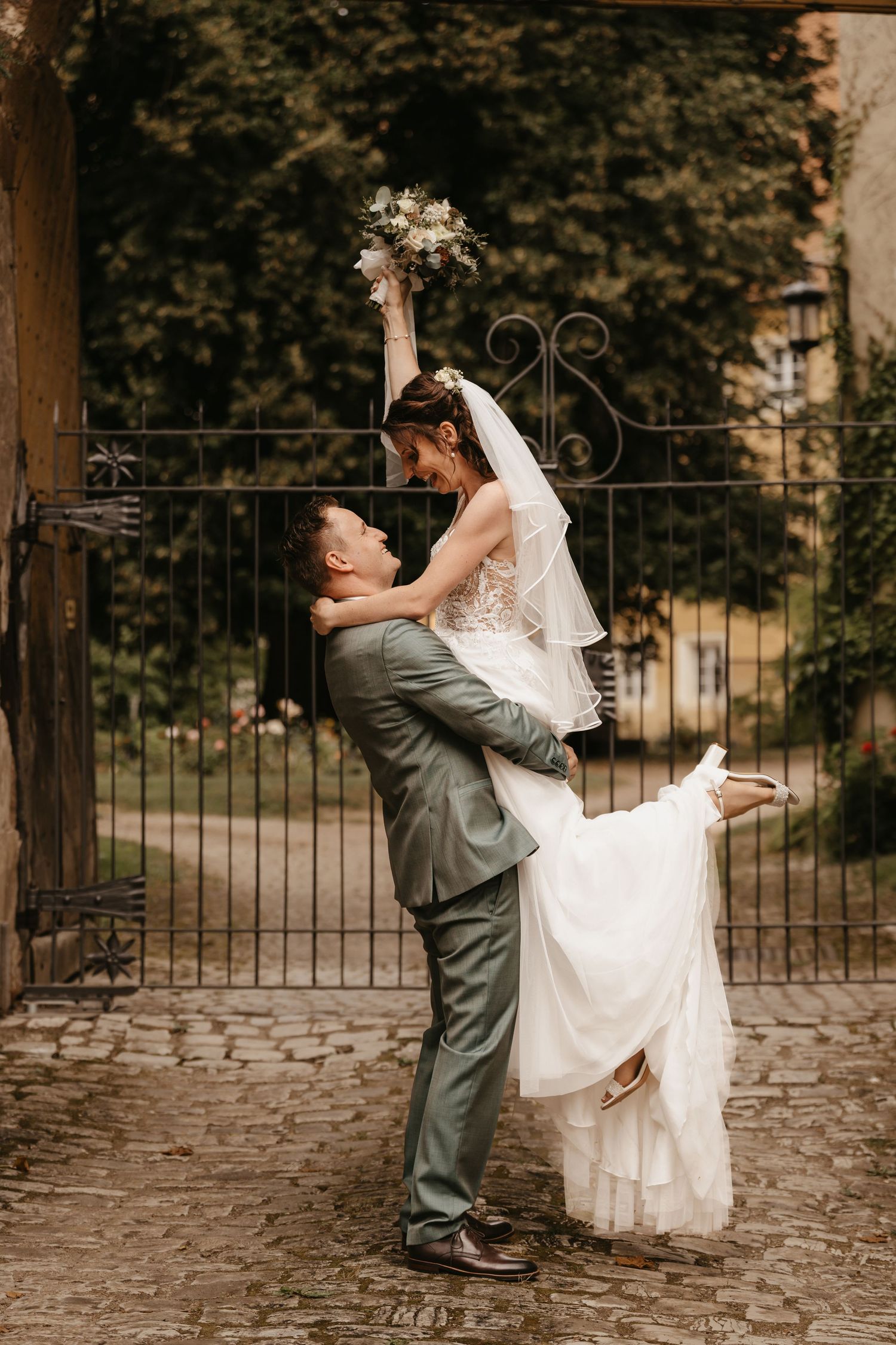 A couple in wedding attire share a playful moment while holding the bride's flowing white dress outdoors.