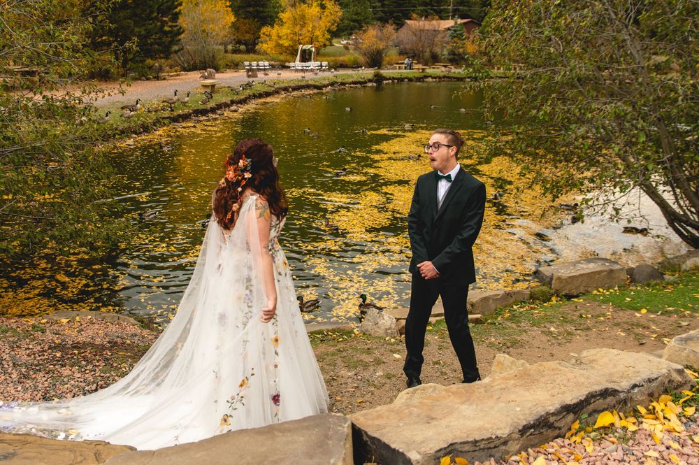 A couple in wedding attire stands by a scenic pond with autumn foliage reflecting in the water.