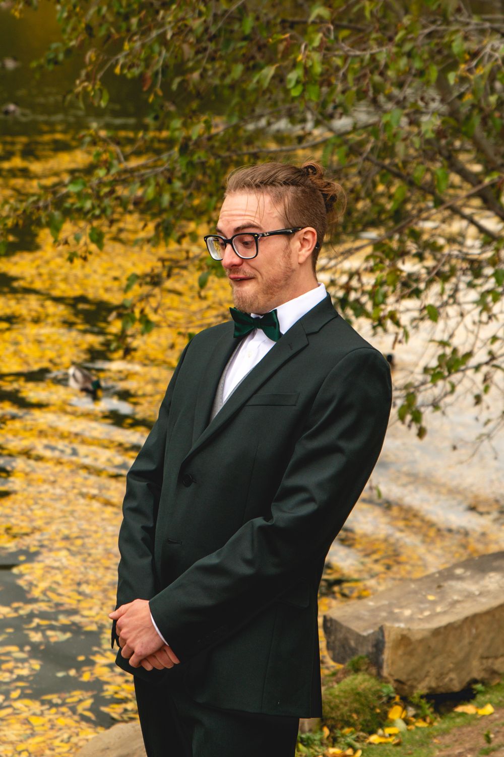 A person in a black suit stands thoughtfully in an autumn setting with yellow fallen leaves in the background.