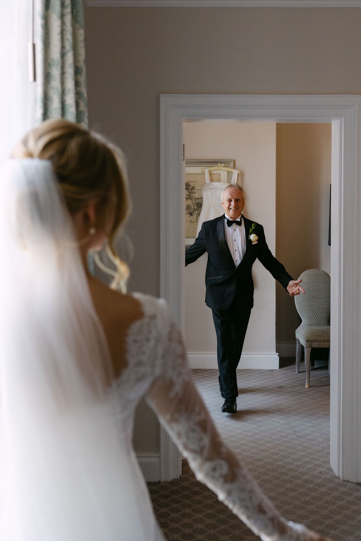 A touching first look moment between a bride in white lace and someone in a dark suit in a bright hallway.