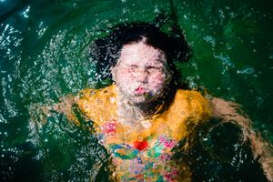 A person wearing a yellow swimsuit splashes in emerald green water creating dynamic water droplets and ripples.