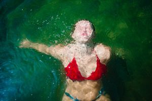 A person wearing a red swimsuit floats on their back in a deep green swimming pool, creating a ethereal underwater scene.