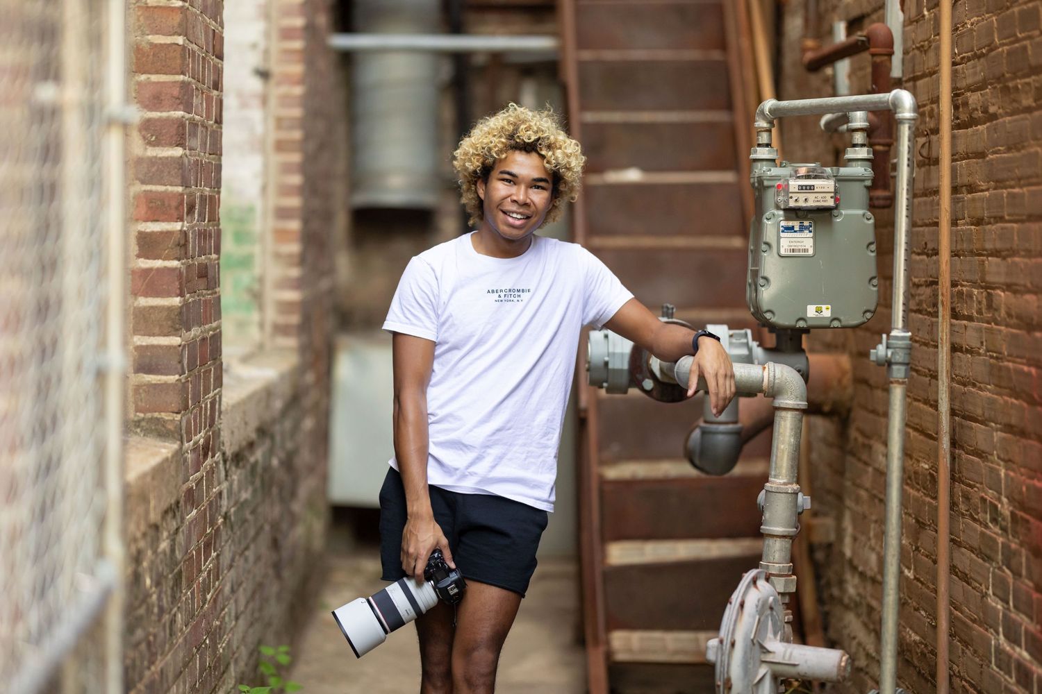Person with blonde curly hair wearing a white t-shirt and black shorts poses in an urban alley holding camera.