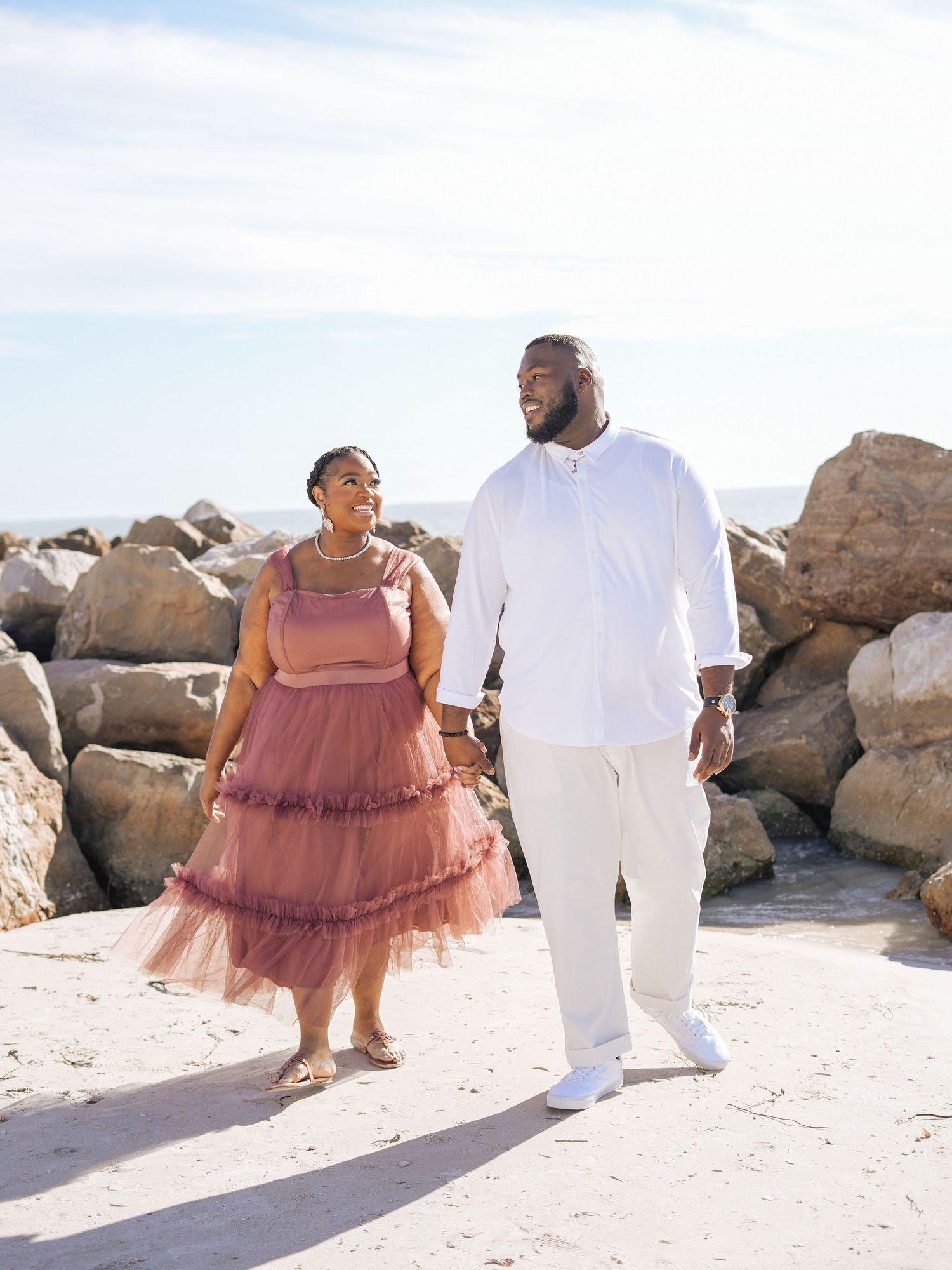 A couple walks together on a sandy beach, wearing coordinated outfits with her in pink tulle dress and him in white attire.
