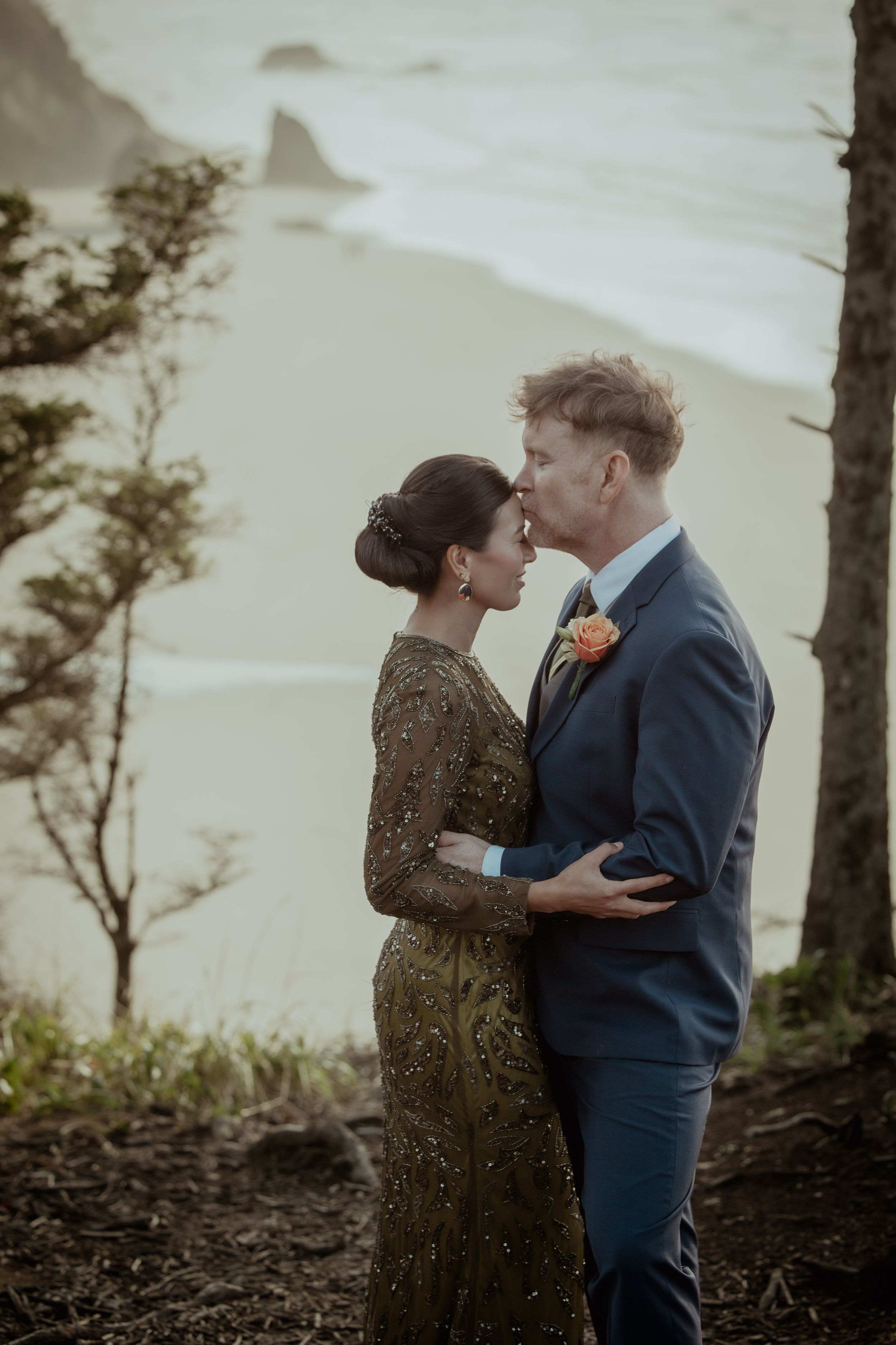 A romantic couple embraces against a misty coastal backdrop with trees silhouetted at sunset.