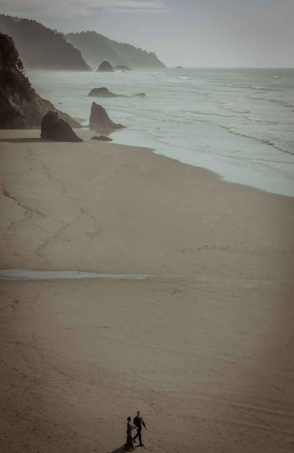 Misty coastal beach landscape with dramatic rock formations and small silhouettes on the sand.