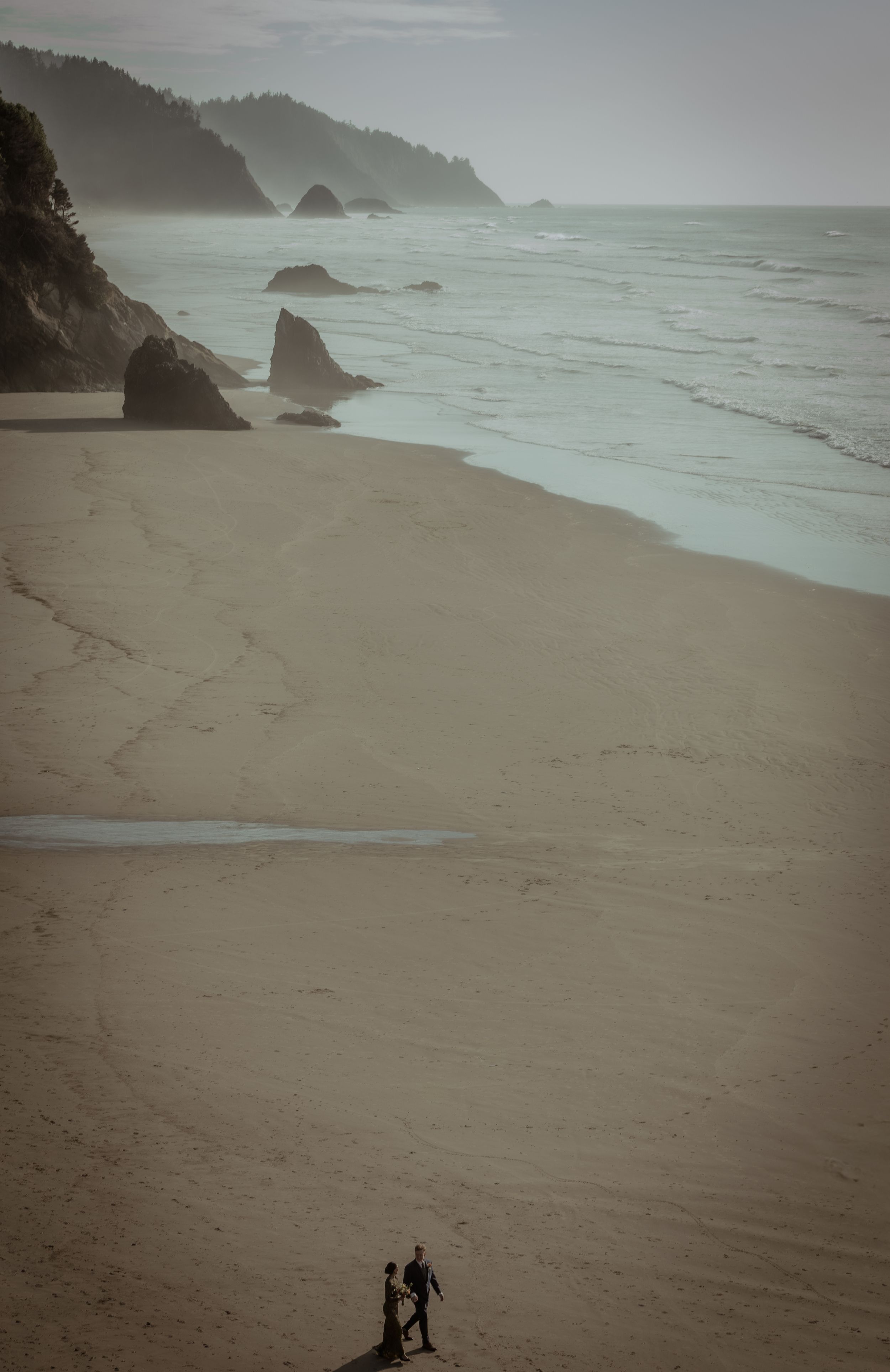 Misty coastal beach landscape with dramatic rock formations and small silhouettes on the sand.
