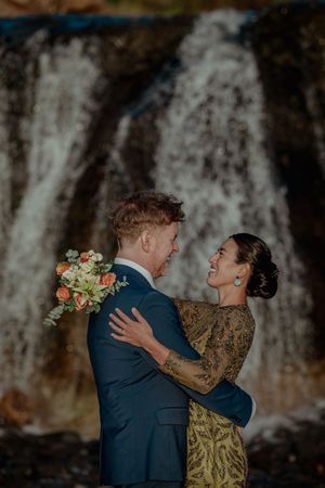 A couple shares an intimate moment in front of a cascading waterfall while holding a floral bouquet.