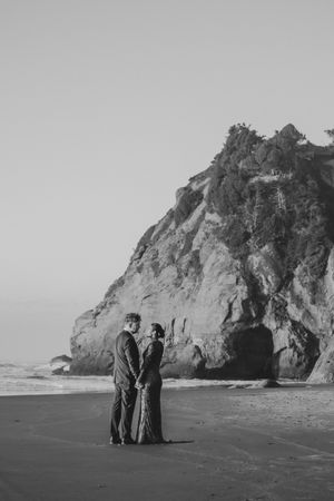 A couple stands together on a scenic beach with dramatic rocky cliffs in the background.