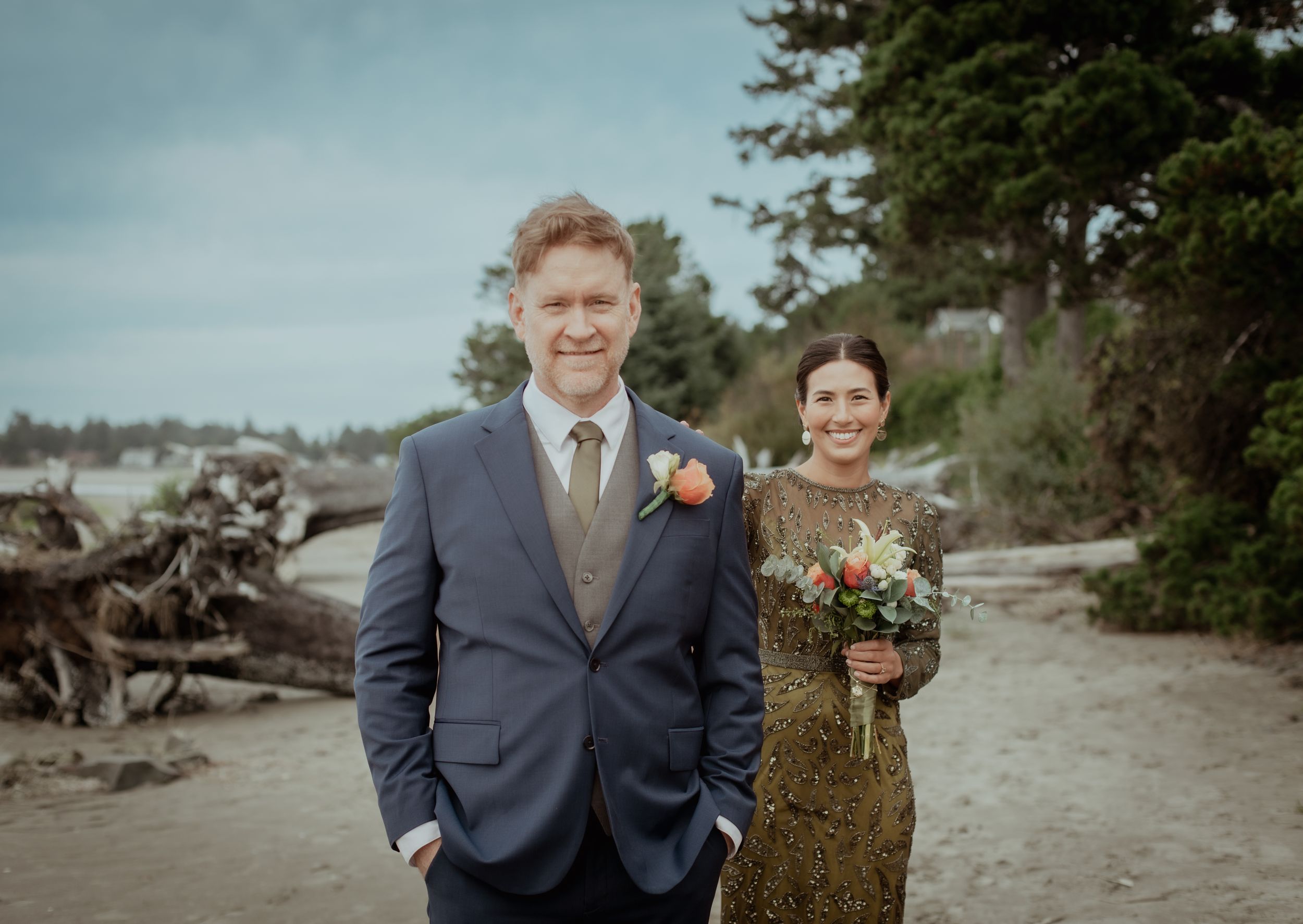 Wedding couple poses on a scenic beach at sunset with mountains and driftwood in the background.