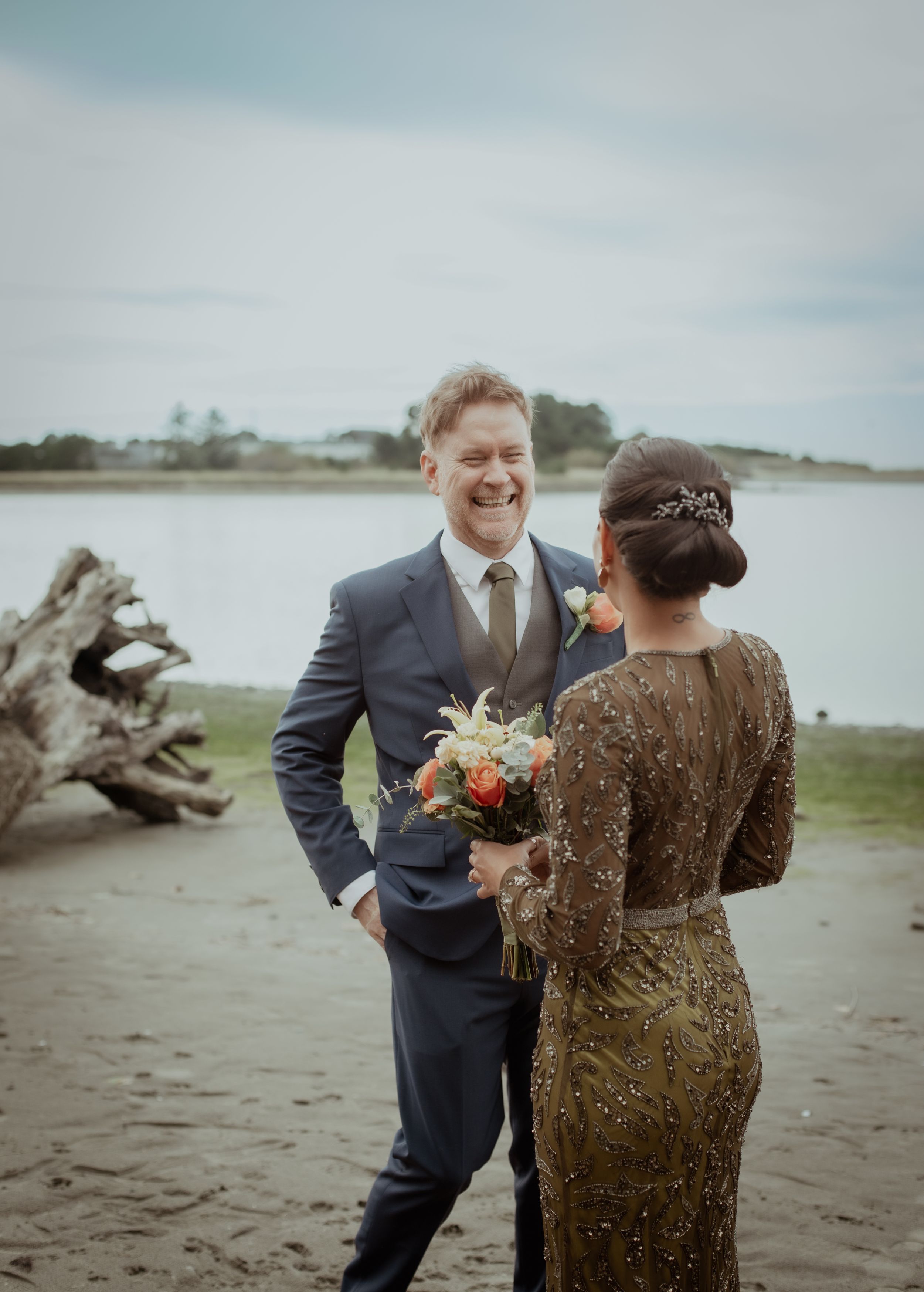 A couple shares a joyful moment together on a beach with driftwood and water in the background.