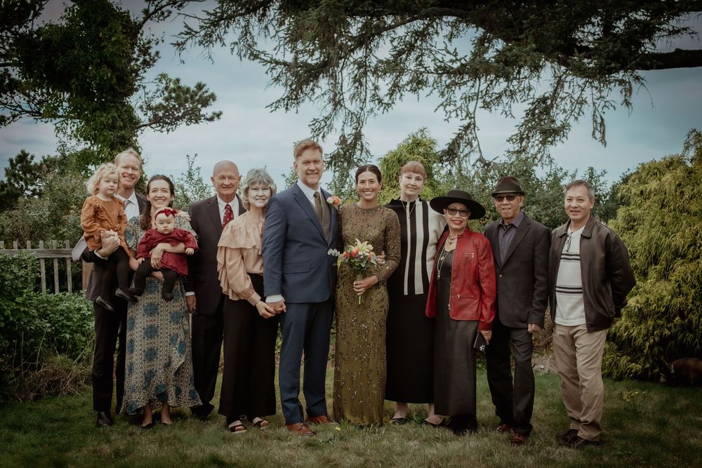 Large group of wedding guests pose together outdoors under trees for a formal group photo.